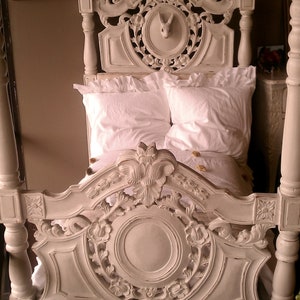 May include: A white, ornate four-poster bed with detailed carvings. The headboard features a circular design with floral accents and a rabbit sculpture. The bed is made up with white linens and pillows, creating a vintage aesthetic.