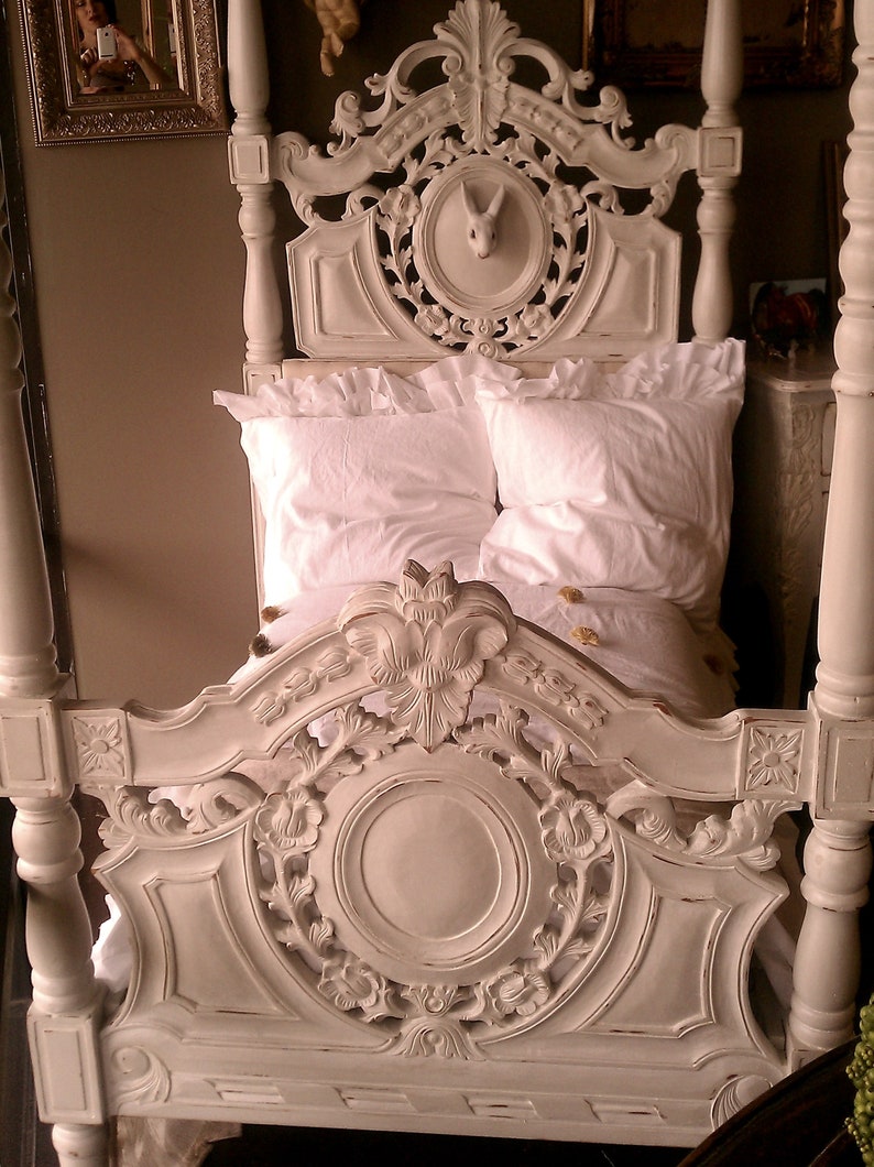 May include: A white, ornate four-poster bed with detailed carvings. The headboard features a circular design with floral accents and a rabbit sculpture. The bed is made up with white linens and pillows, creating a vintage aesthetic.