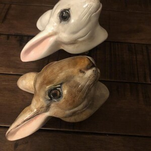 May include: Two ceramic rabbit heads, one white with pink ears and one brown with pink ears. Both have realistic details and painted eyes.
