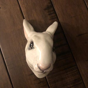 May include: A white ceramic rabbit head with detailed facial features, including dark eyes and a pink nose. The rabbit's ears are upright, and the sculpture is set against a dark wood background.