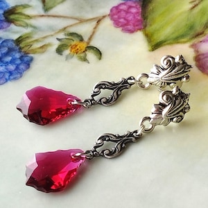 Puede incluir: Un par de pendientes colgantes con un diseño de filigrana plateada y un cristal rojo oscuro en forma de lágrima.