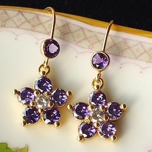 May include: A pair of gold earrings with a flower design. Each earring features a cluster of purple gemstones surrounding a smaller clear gemstone. The earrings dangle from a single purple gemstone.