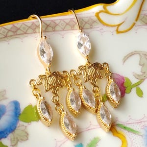 May include: Gold-toned dangle earrings featuring clear, marquise-cut crystals and floral accents. The earrings have a chandelier design with multiple crystal dangles and are suspended from a hook. The earrings are displayed on a decorative, floral-patterned surface.