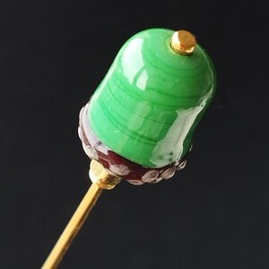 May include: A decorative lapel pin featuring a green and gold design. The top section is a rounded shape with green and white stripes. The base is dark red with gold accents. The pin is mounted on a gold-coloured stem.