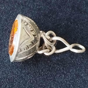 May include: A silver pendant featuring an amber-coloured stone. The pendant has an oval-shaped stone set in a silver frame with detailed engraved patterns. The bail is a looped design. The pendant is shown on a dark blue surface.