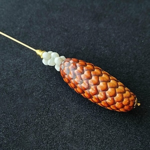 May include: A decorative stick pin featuring a brown, textured pinecone-shaped element attached to a thin gold-toned metal stem. A cluster of small, white, flower-shaped beads is positioned between the pinecone and the metal pin stem.