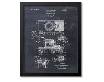 Op de afbeelding: Zwart ingelijste print van een vintage camera patent illustratie. Het witte diagram toont de componenten van de camera, met cijfers en tekst. De print bevat de datum 18 februari 1936 en de naam H. Kappenbender.