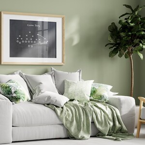Puede incluir: Un sof&aacute; blanco con almohadas y una manta de tiro verdes. El sof&aacute; est&aacute; frente a una pared verde salvia con una pizarra enmarcada con el texto "Corn Dog" y un dibujo de una jugada de f&uacute;tbol. Una planta verde frondosa est&aacute; en la esquina de la habitaci&oacute;n.