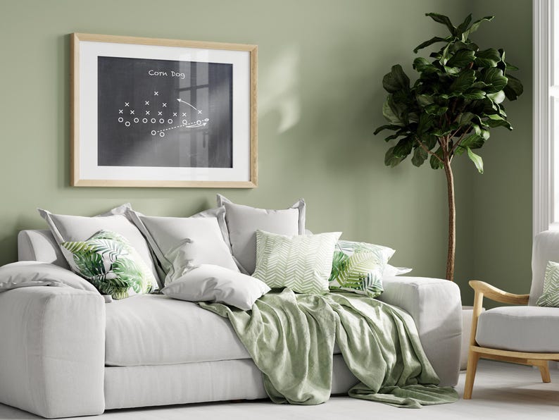 Puede incluir: Un sof&aacute; blanco con almohadas y una manta de tiro verdes. El sof&aacute; est&aacute; frente a una pared verde salvia con una pizarra enmarcada con el texto "Corn Dog" y un dibujo de una jugada de f&uacute;tbol. Una planta verde frondosa est&aacute; en la esquina de la habitaci&oacute;n.