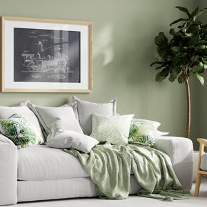 May include: A white sofa with a green throw blanket and cushions featuring a tropical leaf design. A framed black and white print of a train engine diagram hangs on the wall behind the sofa. A green leafy plant is in the corner of the room.