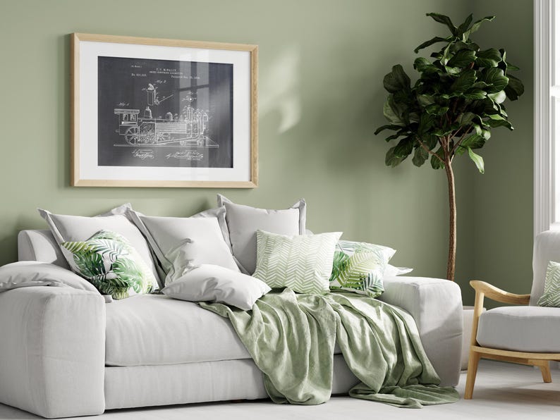 May include: A white sofa with a green throw blanket and cushions featuring a tropical leaf design. A framed black and white print of a train engine diagram hangs on the wall behind the sofa. A green leafy plant is in the corner of the room.
