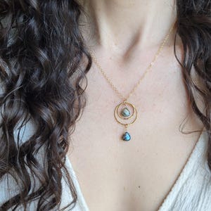 May include: Gold necklace with a double-circle pendant holding a grey stone and a blue teardrop. The chain is gold. The jewellery is shown against a white top. The necklace is a delicate piece.