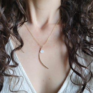 May include: A gold-toned necklace featuring a delicate chain and a pendant composed of a small, translucent, teardrop-shaped stone attached to a curved, crescent-shaped metal bar with a textured, hammered finish.