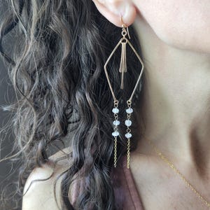 May include: Gold-tone geometric dangle earrings. The earrings feature a diamond-shaped frame with vertical bars and small, light-colored stones suspended from delicate chains. The earrings are shown on a person's ear.