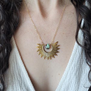 May include: A gold necklace with a sunburst pendant and a teardrop-shaped green gemstone. The necklace is worn by a person with long, curly hair.