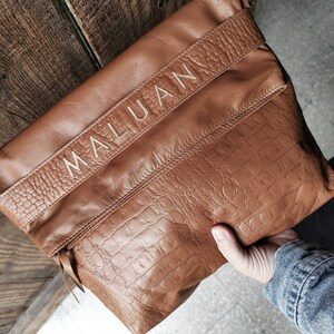 Peut inclure: Un sac à bandoulière en cuir marron avec une fermeture à rabat et une poche zippée. Le sac a une texture en relief de crocodile et le mot "MALUAN" est cousu en or sur le devant.