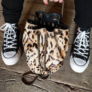 Puede incluir: Un pequeño bolso de cubo de estampado de leopardo con una correa de cadena dorada. El bolso está abierto y el forro es de rayas blancas y negras. El bolso lo lleva una persona que lleva pantalones vaqueros negros y zapatillas negras y blancas.