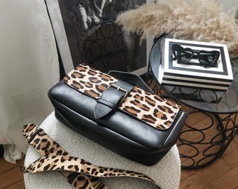 May include: A black leather handbag with a leopard print flap and strap. The bag has a gold buckle closure and is sitting on a white fuzzy surface.