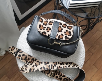 May include: A black leather handbag with a leopard print flap and a gold chain strap. The bag is sitting on a white fuzzy surface with a leopard print strap.