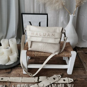 Peut inclure: Un sac à bandoulière en cuir blanc avec le mot "MALUAN" en relief sur le rabat avant. Le sac est posé sur un tabouret en bois blanc avec un siège en osier. Il y a deux ceintures en cuir blanc avec des franges sur le sol devant le tabouret.