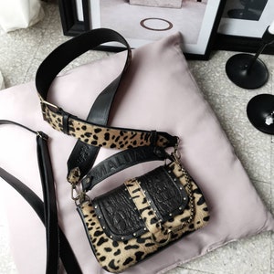 Può includere: Una piccola borsa a tracolla in stampa leopardata nera e marrone con una tracolla in pelle nera e un dettaglio a catena dorata. La borsa ha una chiusura a lembo e una piccola tasca sul davanti. La borsa è appoggiata su un cuscino rosa.