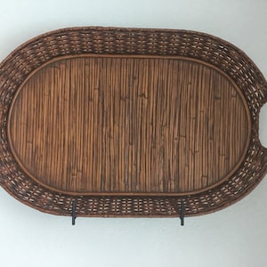 May include: Oval woven wicker tray with integrated handles. The tray is brown with a natural texture and is supported by two black metal brackets. The tray is designed for serving or decorative purposes.