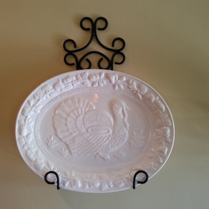 May include: White ceramic serving platter with a raised design of a turkey. The platter is hanging on a black metal wall mount.