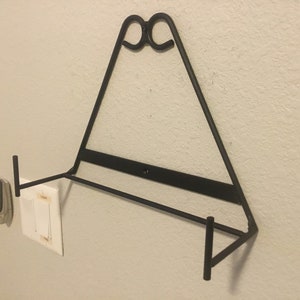 May include: Black metal wall-mounted shelf with a triangular design and two horizontal bars. The shelf is mounted on a white wall.