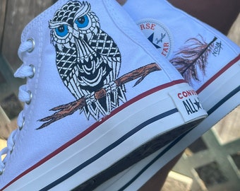Könnte beinhalten: Weiße High-Top-Canvas-Sneaker mit handgemaltem Design. Ein Schuh zeigt eine Eule mit blauen Augen und schwarz-weißen Federn. Der andere Schuh zeigt eine braune Feder mit dem Text "Nova" in Schwarz.