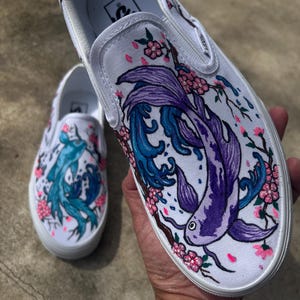 Op de afbeelding: Een paar witte canvas slip-on schoenen met handgeschilderde ontwerpen. De ene schoen heeft een paarse en blauwe koi-vis die door een kersenbloesemtak zwemt. De andere schoen heeft een blauwe koi-vis die door een kersenbloesemtak zwemt.