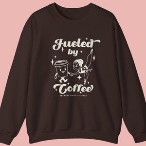 May include: A dark brown sweatshirt with a white graphic that reads "Fueled by Coffee" with a cartoon coffee cup and ballerina. The text "BECAUSE BALLET IS HARD" is below the main text.