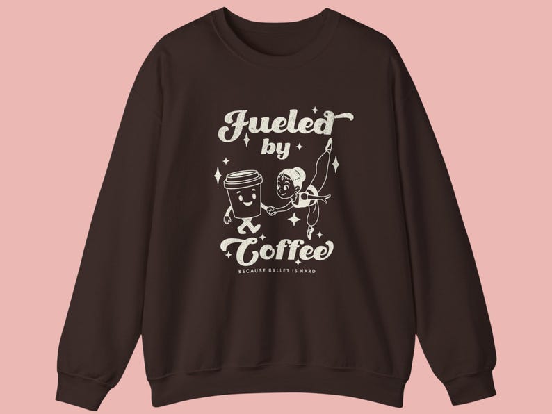 May include: A dark brown sweatshirt with a white graphic that reads "Fueled by Coffee" with a cartoon coffee cup and ballerina. The text "BECAUSE BALLET IS HARD" is below the main text.