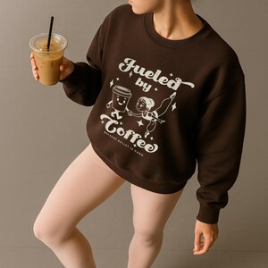 May include: A brown sweatshirt with the text "Fueled by Coffee" and a graphic of a coffee cup and a ballerina. The sweatshirt is worn with pink tights and ballet shoes. A person is holding a cup of iced coffee.
