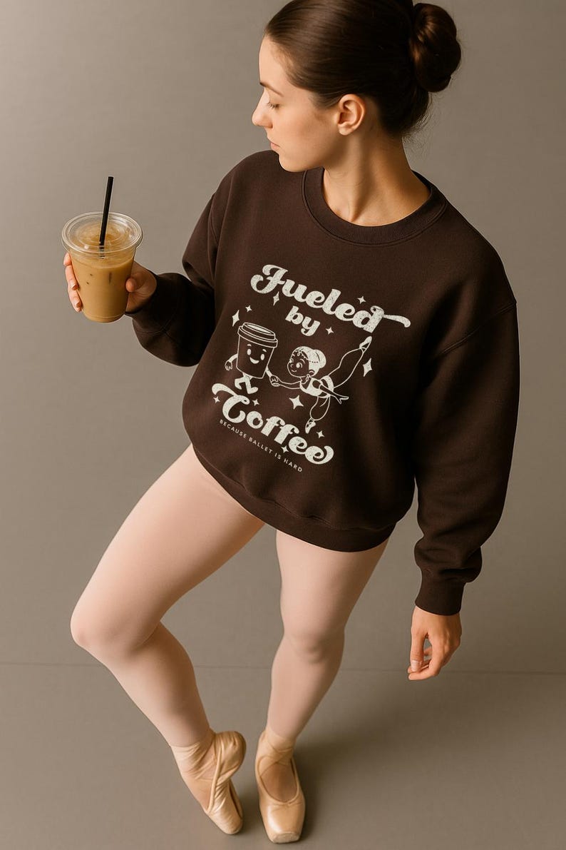May include: A brown sweatshirt with the text "Fueled by Coffee" and a graphic of a coffee cup and a ballerina. The sweatshirt is worn with pink tights and ballet shoes. A person is holding a cup of iced coffee.