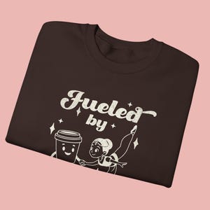 May include: A dark brown t-shirt with the words "Fueled by" above a cartoon coffee cup and a ballerina. The graphic is in white. The shirt is displayed on a pink background.