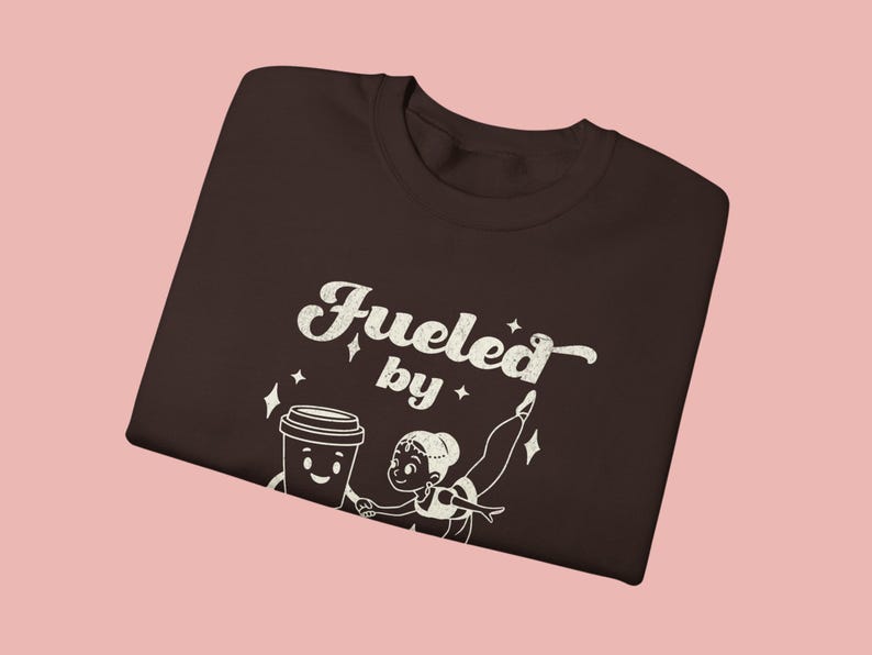 May include: A dark brown t-shirt with the words "Fueled by" above a cartoon coffee cup and a ballerina. The graphic is in white. The shirt is displayed on a pink background.