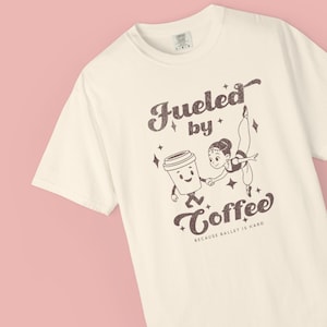 May include: Off-white t-shirt with a graphic of a coffee cup and a ballerina. The text says "Fueled by Coffee" and "Because Ballet is Hard" in a retro font. The shirt is set against a pink backdrop.