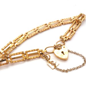 May include: A gold-toned bracelet featuring a series of rectangular, textured links connected by a heart-shaped padlock charm. A small, thin chain attaches the padlock to the bracelet, and a small rectangular tag hangs near the clasp mechanism.