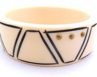May include: A cream-colored bangle bracelet featuring a repeating geometric pattern of dark brown etched lines. The design includes triangular shapes with three small, circular, metallic gold-toned studs embedded within the center of each triangle.