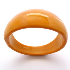 May include: A wide, amber-coloured bangle bracelet. The smooth, polished surface reflects light, creating highlights. The bracelet is a solid, rounded shape, with a hollow interior. The colour is a warm, translucent orange-brown.