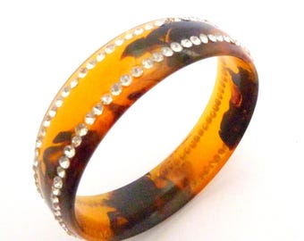 May include: A circular bangle bracelet featuring a translucent amber and dark brown tortoiseshell pattern. The bracelet is accented with two parallel rows of small, clear, reflective rhinestones that encircle the entire outer surface.