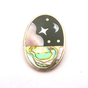 May include: An oval-shaped brooch featuring a celestial inlay design. The upper section displays a black background with a white mother-of-pearl crescent moon and star, alongside small gold-tone dots. The lower section features iridescent abalone shell.