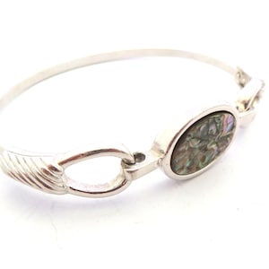 May include: A silver-toned bracelet with an oval abalone shell centerpiece. The bracelet features a hinged design with a textured detail on one side. The abalone shell has iridescent colors of green, blue, and purple.