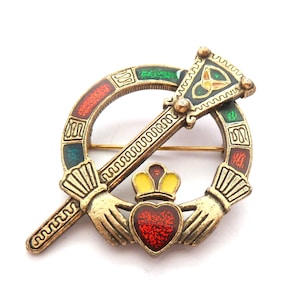 May include: A gold-toned brooch featuring a Claddagh design with a heart, crown, and hands. The brooch has a circular frame with red, green, and blue enamel accents. A decorative bar crosses the circle, and the brooch is set against a white background.