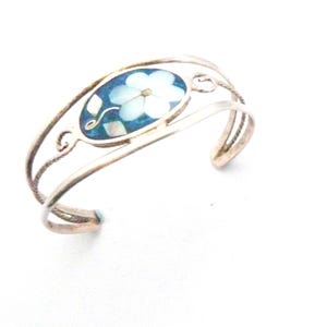 May include: Silver-toned cuff bracelet featuring an oval centerpiece with a blue background and a white flower design. The bracelet has an open design with decorative swirls and a polished finish. A delicate piece of jewelry.