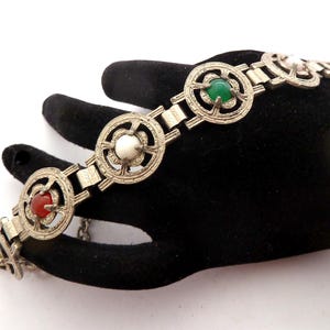 Puede incluir: Pulsera plateada con eslabones circulares y patrones decorativos. Cada eslabón contiene una piedra preciosa en rojo, verde o blanco. La pulsera se muestra sobre una superficie negra, resaltando su intrincado diseño y atractivo vintage.