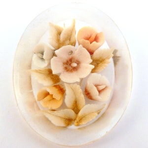 May include: A white oval cameo brooch featuring a raised design of flowers in shades of white, peach, and brown.