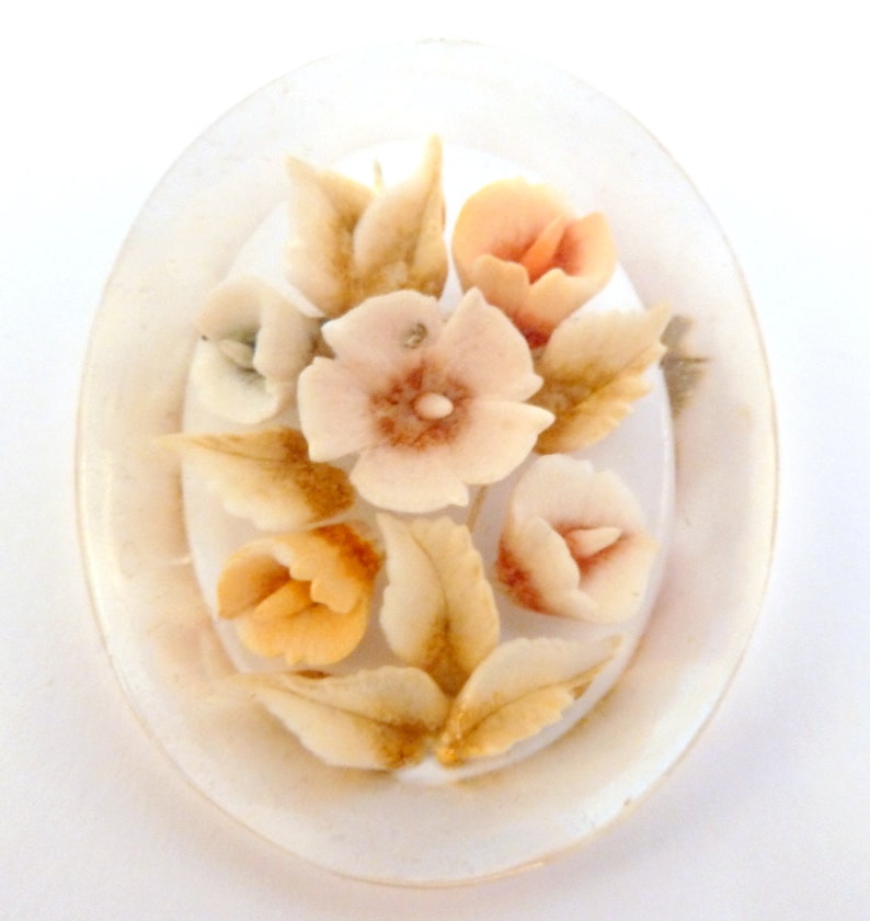 May include: A white oval cameo brooch featuring a raised design of flowers in shades of white, peach, and brown.