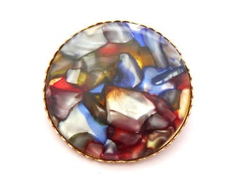 Könnte beinhalten: Eine runde Brosche mit goldfarbenem Rand. Die Mitte der Brosche zeigt ein Mosaikmuster aus bunten, unregelmäßigen Formen in Rot, Blau, Weiß und Gelb. Die Brosche ist vor einem weißen Hintergrund platziert.