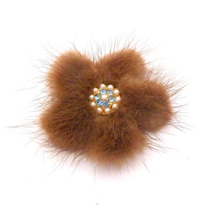 May include: A brown fur flower brooch with a gold-tone center. The center of the flower is decorated with small, light blue rhinestones and faux pearls. The brooch has a pin backing for secure attachment to clothing or accessories.